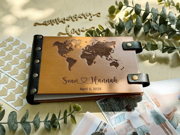 Wooden photo album with world map design and engraved names, surrounded by greenery and photos.